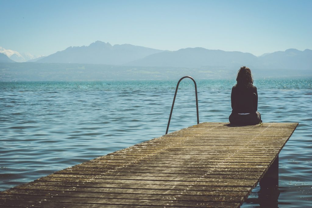 a woman sits on the end of a dock during daytime staring across a lake
